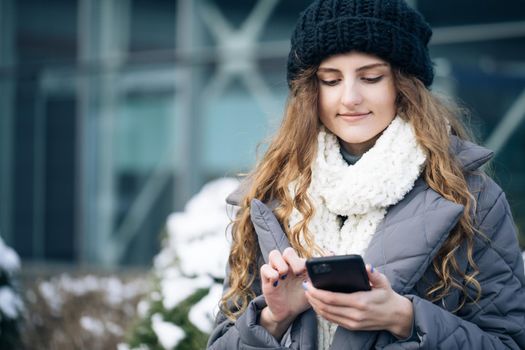 Female using smartphone standing outside. Smiling happy girl employee typing on a cellphone. Joyful young female in good mood typing and scrolling on smartphone outdoors. Winter concept.