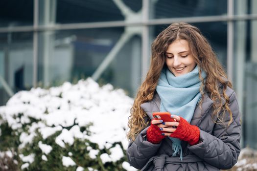 Curly-haired female texting on smartphone standing on street in winter city. Female tapping on cellphone outdoors. Vacation winter outdoor. Happy Young Woman Enjoys Life