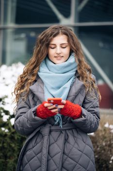 Joyful young female in good mood typing and scrolling on smartphone outdoors. Curly-haired female using smartphone standing outside. A smiling happy girl employee typing on a cellphone. Winter concept