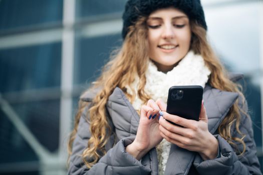 Joyful young female in good mood typing and scrolling on smartphone outdoors. Winter concept. Female using smartphone standing outside. Happy girl employee typing on a cellphone.