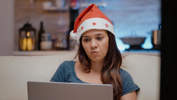 Festive person working with laptop and watching television on christmas holiday. Woman with santa hat looking at device and TV screen while eating chips on winter celebration