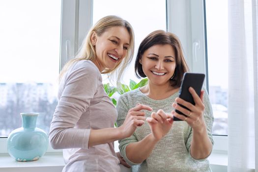 Two middle-aged women friends looking together at smartphone screen. Females at home near window in winter season. Relationship, lifestyle, leisure, friendship, technology, mature people concept