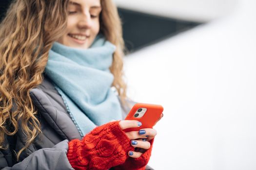 Portrait of cheerful Curly-haired Young female texting on smartphone standing on street in winter city on New Year. Female tapping on cellphone outdoors. Online shopping, buying new year's gifts