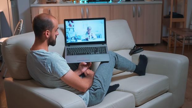 Doctor consulting sick patient during coronavirus quarantine using laptop computer for videocall consultation sitting on sofa. Caucasian man disscusing about illness symptoms and headlcare treatment