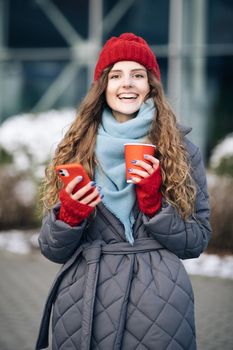 Female look around on the street city. Modern stylish curly woman with cup coffee, uses her smartphone . Being online, social networks.