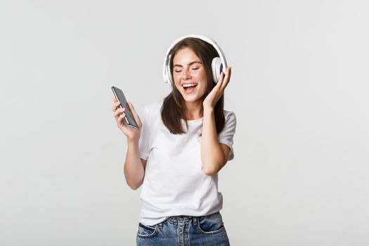 Happy beautiful brunette girl dancing and listening music in wireless headphones, holding smartphone.