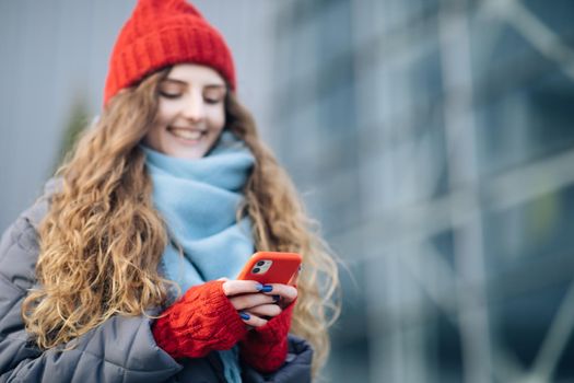 Joyful female in good mood typing and scrolling on smartphone outdoors. Curly-haired female using smartphone standing outside. A smiling happy girl employee typing on a cellphone. Winter concept
