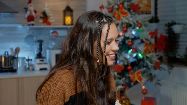 Young woman using modern technology for video call communication with friends in festive kitchen. Caucasian person making preparations on laptop for christmas day dinner celebration