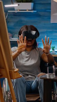 Black artist woman using vr glasses for art project in workshop room at home. African american person with handicap sitting in wheelchair while having headset for masterpiece