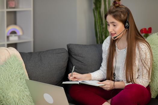 Smiling teenage girl wearing headphones using laptop sitting on bed. Happy teen school student conference calling on computer for online distance learning communicating with friend by webcam at home.