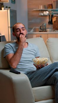 Man eating popcorn and watching TV, enjoying mid-night snack. Relaxed man lying on sofa late at night while watching entertainment tv shows at home cinema eating popcorn