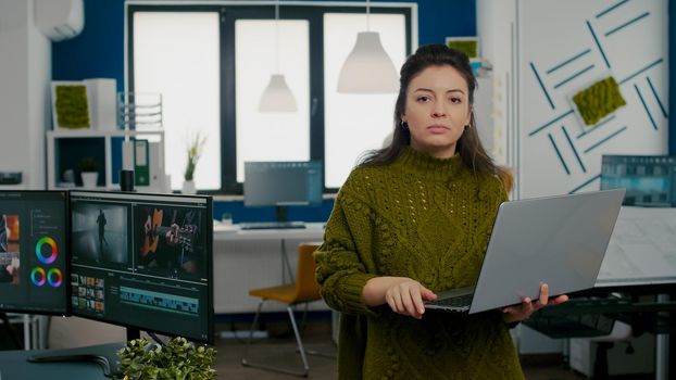 Employee looking at camera smiling standing in front of camera working in creative agency office holding laptop. Videographer works in multimedia studio production editing video in modern workplace.