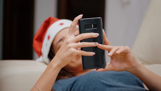 Close up of festive woman looking at smartphone and TV for christmas eve celebration. Person holding gadget and watching television while laying on couch on winter holiday. Adult with device