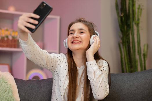 Pretty smiling girl taking selfie on smartphone, listening to music, sitting on couch at home. Beautiful woman listening to music at home