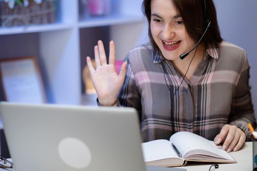 Happy woman wearing headphones greeting during a video conference on line with a red laptop sitting in a table in the living room.