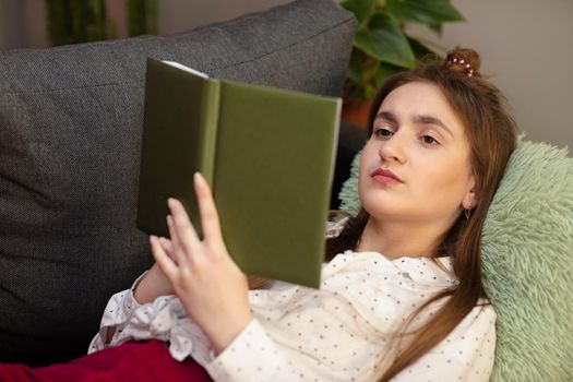 Teenager girl home - student read book laying on sofa. Portrait of smiling woman holding book and lying on couch.