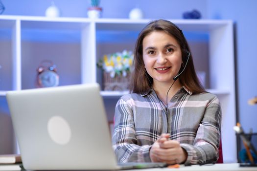 Positive charming woman working at home. Smiling young woman student wear wireless headset using laptop for e learning course online teaching in internet at home