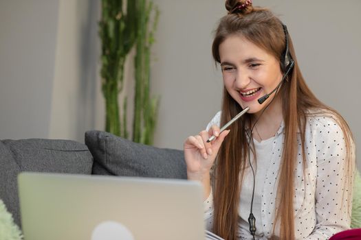 Smiling teen girl wearing headphones listening to audio course using laptop at home, making notes, young woman learning foreign languages, digital self education, studying online, enjoying music