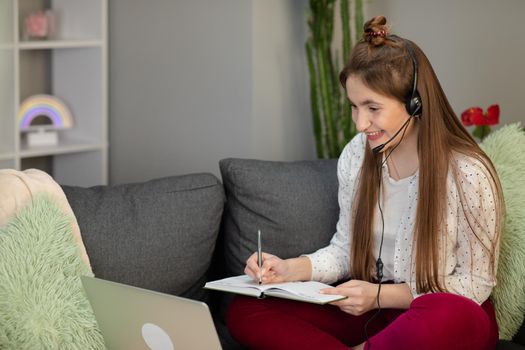 Teenage girl wearing headphones using laptop sitting on bed. Happy teen school student conference calling on computer for online distance learning communicating with friend by webcam at home