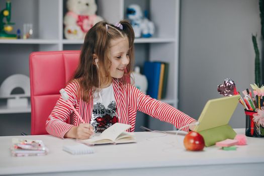 Young preschool girl watching lesson online and studying from home. Kid girl taking notes while looking at computer screen following professor doing math on video call.