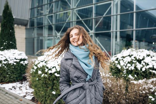 Charming young curly woman with a magnificent brown hair, big brown eyes and stylish look turns to camera and smiles. Attractive young lady is rushing in the winter city-center.