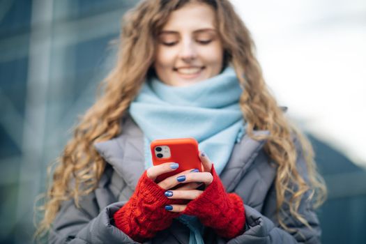 Curly-haired female using smartphone standing outside. A smiling happy girl employee typing on a cellphone. Winter concept. Joyful young female in good mood typing and scrolling on smartphone outdoors