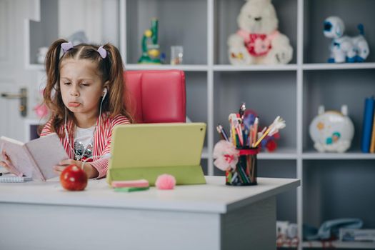 Kid girl make video call tutoring write notes, teaching concept. Preschool girl watching lesson online and studying from home. Kid girl taking notes while looking at computer screen