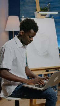 African american artist man searching inspiration on laptop computer sitting in studio space. Black young creative adult working on drawing with online technology for next masterpiece