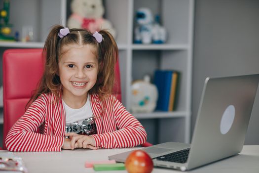 Portrait of a preschool girl looking at the camera and smiling. Kid girl sitting at the desk in children room at home and studying online. Distance learning, school online.