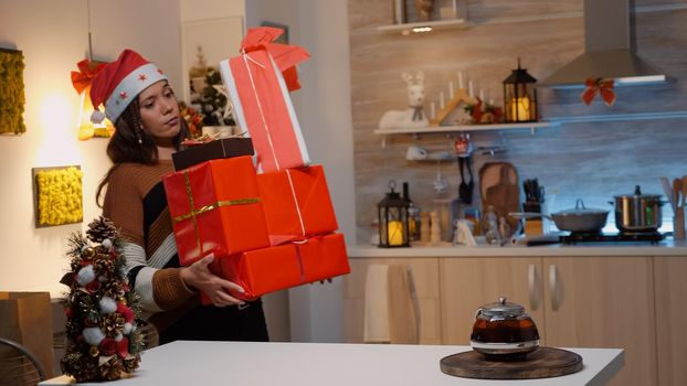 Festive woman bringing boxes of gifts on kitchen counter at holiday decorated home. Young caucasian adult preparing presents with wrapping paper for friends and family at christmas eve party