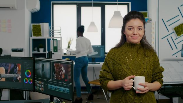 Portrait of creative designer smiling at camera holding a cup of coffee standing in start up agency office. Woman working in multimedia studio production editing video in workplace with diverse team.