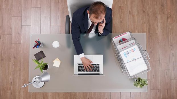 Top view of businessman discussing business profit with partner at phone while typing company statistics on laptop. Executive director working at management strategy in startup corporate office