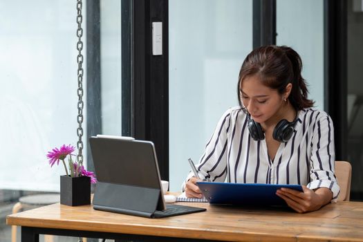 Asian woman taking notes in notebook and using laptop computer for e-learning at home.