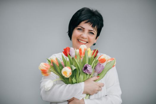 Closeup portrait of elderly woman holding flower bouquet in her hands. Model posing isolated over grey studio background. Charming female 8 march concept.