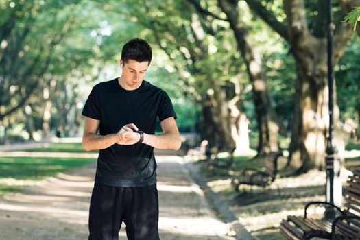 Distance application. Sporty man running along empty road in park checking smartwatch notification online using wireless earphones while training outside. Healthy active lifestyle