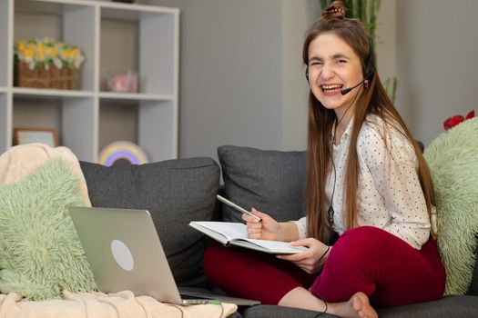 Happy teen school student conference calling on computer for online distance learning communicating with friend by webcam at home. Teenage girl wearing headphones using laptop sitting on bed.