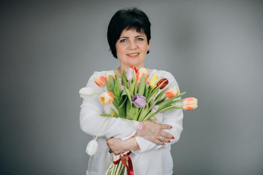 Attractive woman holds bouquet of flowers tulips in her hands gift soft toy looking at camera and smiling. Woman with tulip bouquet. Woman with gifts