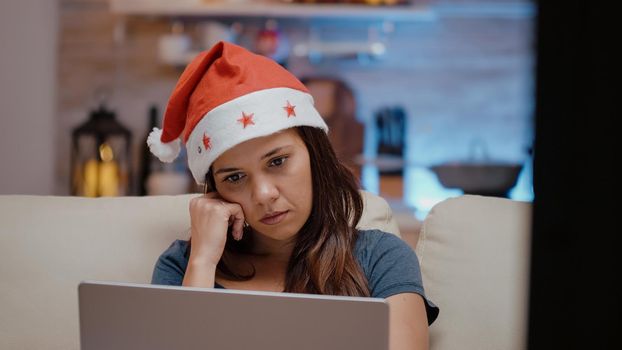 Close up of sleepy adult looking at laptop on christmas eve. Festive woman with santa hat falling asleep on couch while working with technology on device. Tired person on holiday