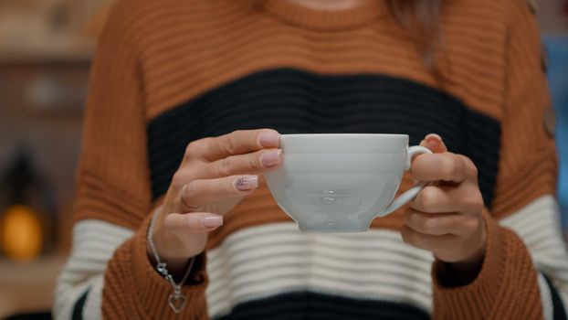 Close up of tea cup held by young woman at home in kitchen decorated with tree and ornaments. Adult preparing to drink from hot mug while waiting on christmas eve holiday dinner party