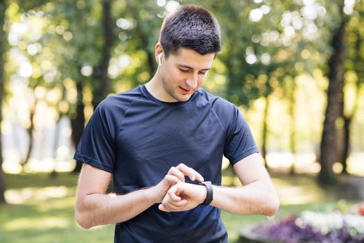 Adult man using smart watch tapping touchscreen enjoying healthy active lifestyle. Male runner working out cardio exercise on summer park. Portable modern earphones.