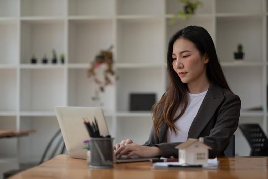 Real estate agent hand typing laptop keyboard with house model and keys, business asian woman working at home office