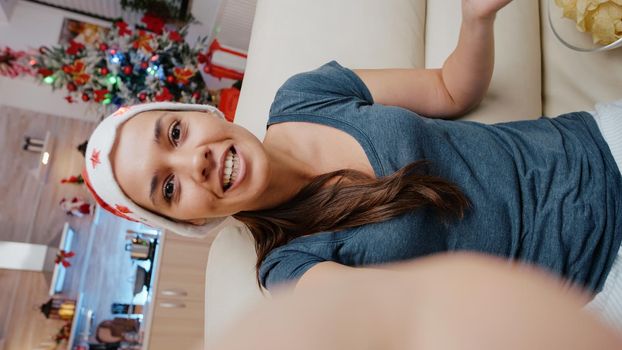 POV of cheerful woman waving at video call conference for christmas celebration with family. Adult using internet and online remote communication for holiday festivity in decorated kitchen.