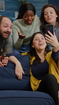 Multiracial friends speaking with collegue man during video call conference using modern smartphone. Group of multi-ethnic people hanging out together, drinking beer, having fun during night party