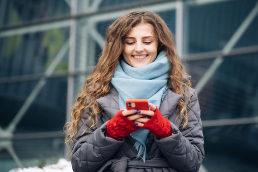 Portrait of cheerful Curly-haired Young female texting on smartphone standing on street in winter city on New Year. Female tapping on cellphone outdoors. Online shopping