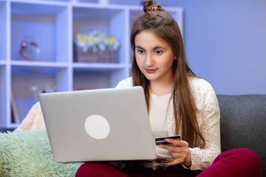 Girl connecting with a laptop and doing online shopping using a credit card. Young girl are buying online with credit card while sitting on sofa. Woman are using laptop and doing online transactions.