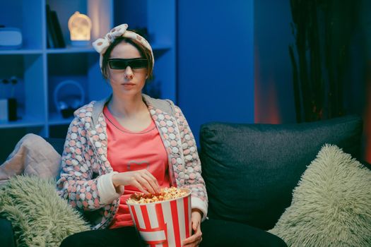 Woman on a soft couch eating popcorn, looking at the TV screen in 3D glasses. Woman eating popcorn in 3d glasses.