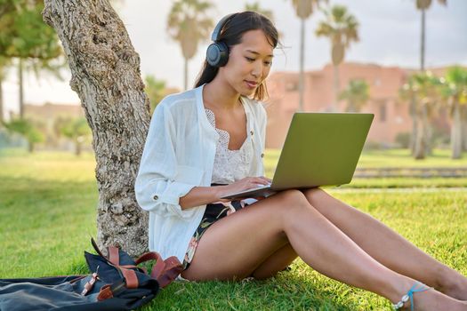 Young woman in headphones with laptop sitting on grass in park. Female student, creative freelancer, businesswoman, blogger working, studying remotely. Education, online business blogging technologies