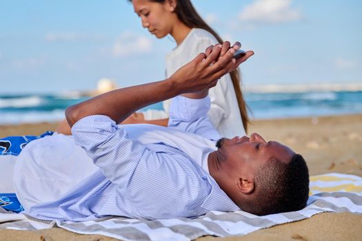 Young beautiful multiethnic couple having a rest on the beach, African American man with a smartphone in his hands close-up. Lifestyle, technology, leisure, people concept