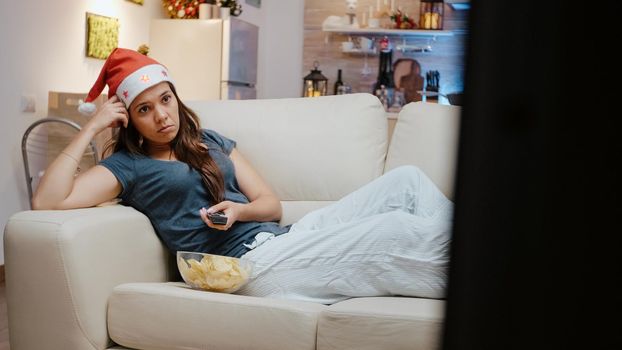 Bored adult holding TV remote control and switching channels on christmas eve. Lonely woman with santa hat looking at movie on television and having bowl with chips on couch.