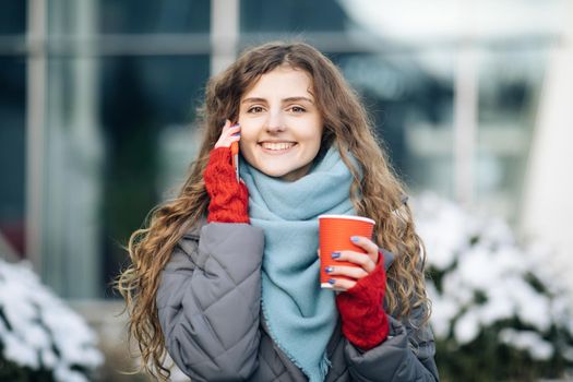 Young curly girl talking on her mobile phone while walking in the city street. Curly haired woman using her smartphone and holding red paper cup with coffee. Woman walking and talking
